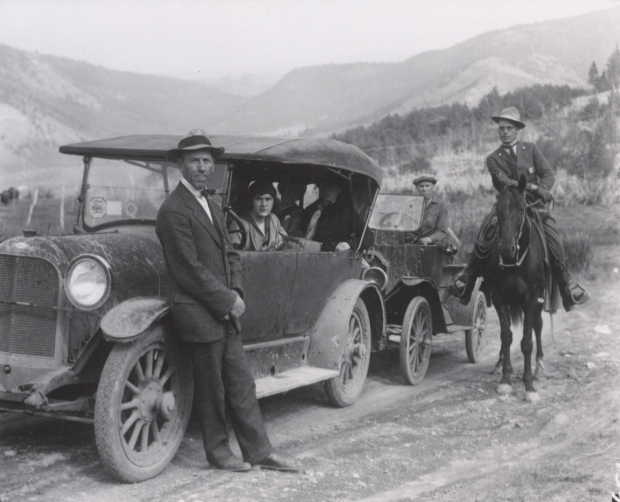 Man in NPS uniform, broad brim hat and a shield shaped badge rides a horse along side visitors in old cars stopped on the side of the road.